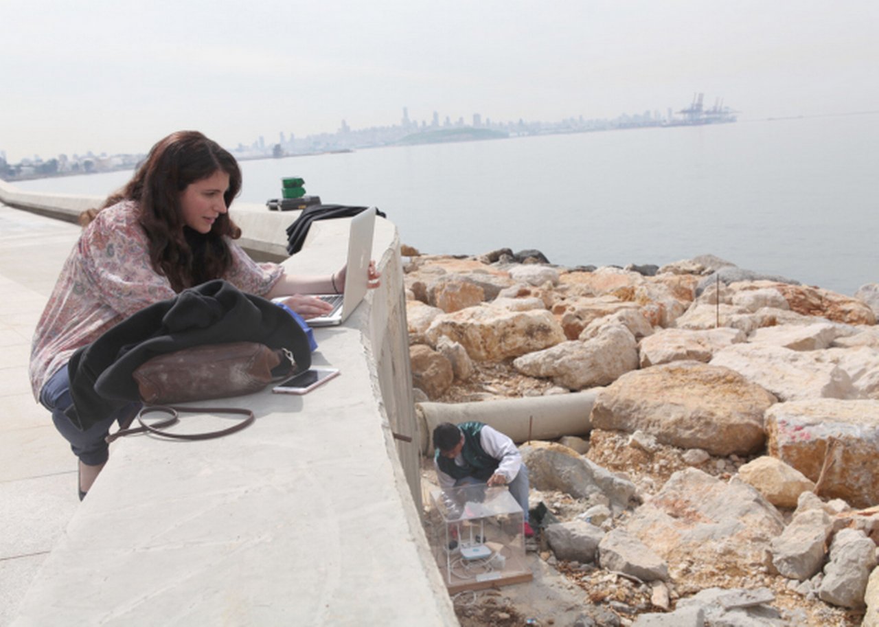 Caline Aoun on the laptop outside with view on rocky seaside